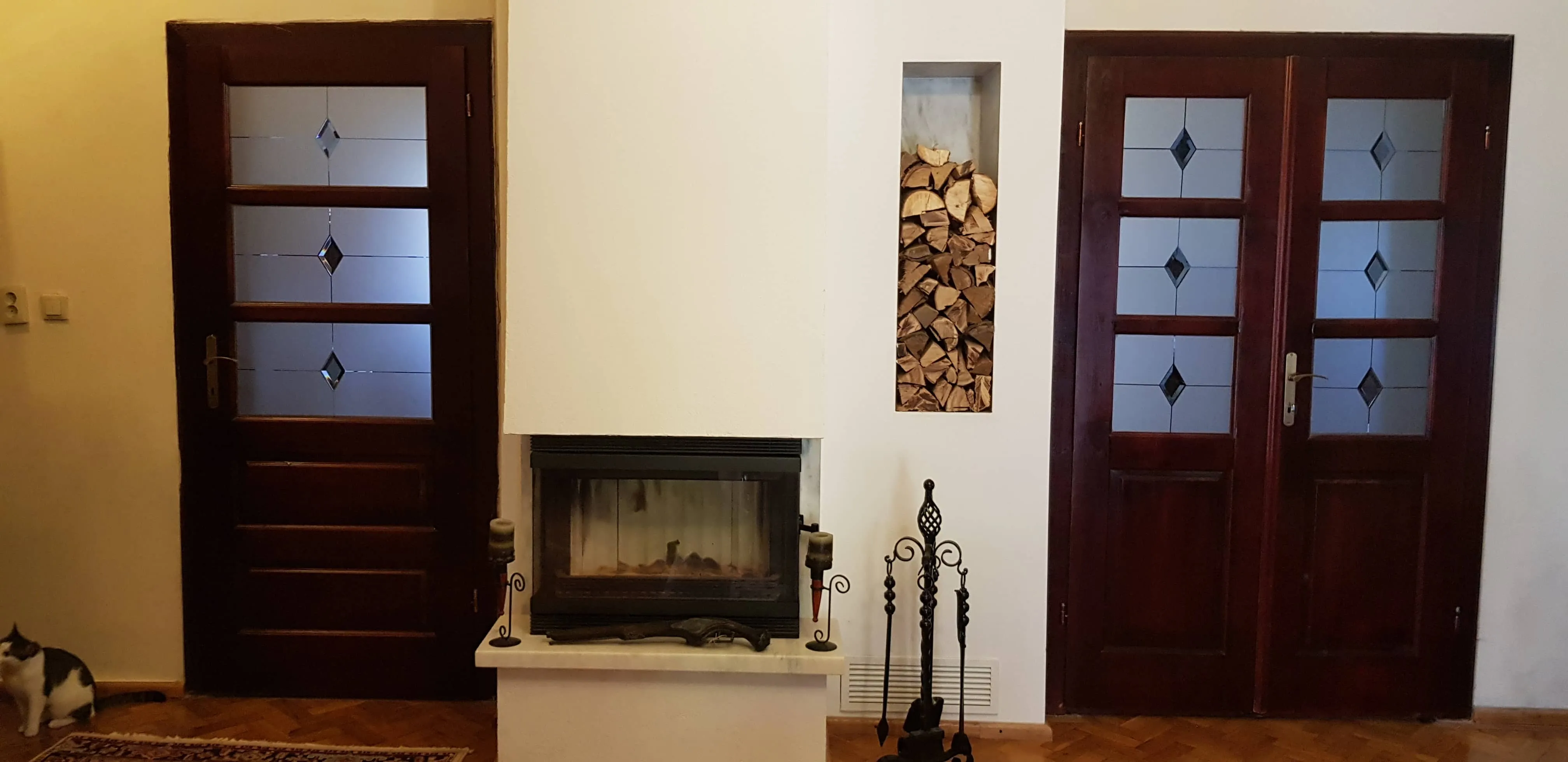 Indoor scene with a white fireplace in the center, flanked by two dark wood doors with frosted glass panels and diamond-shaped designs; a niche filled with stacked firewood is above fireplace tools, and a black and white cat sits on the floor to the left.