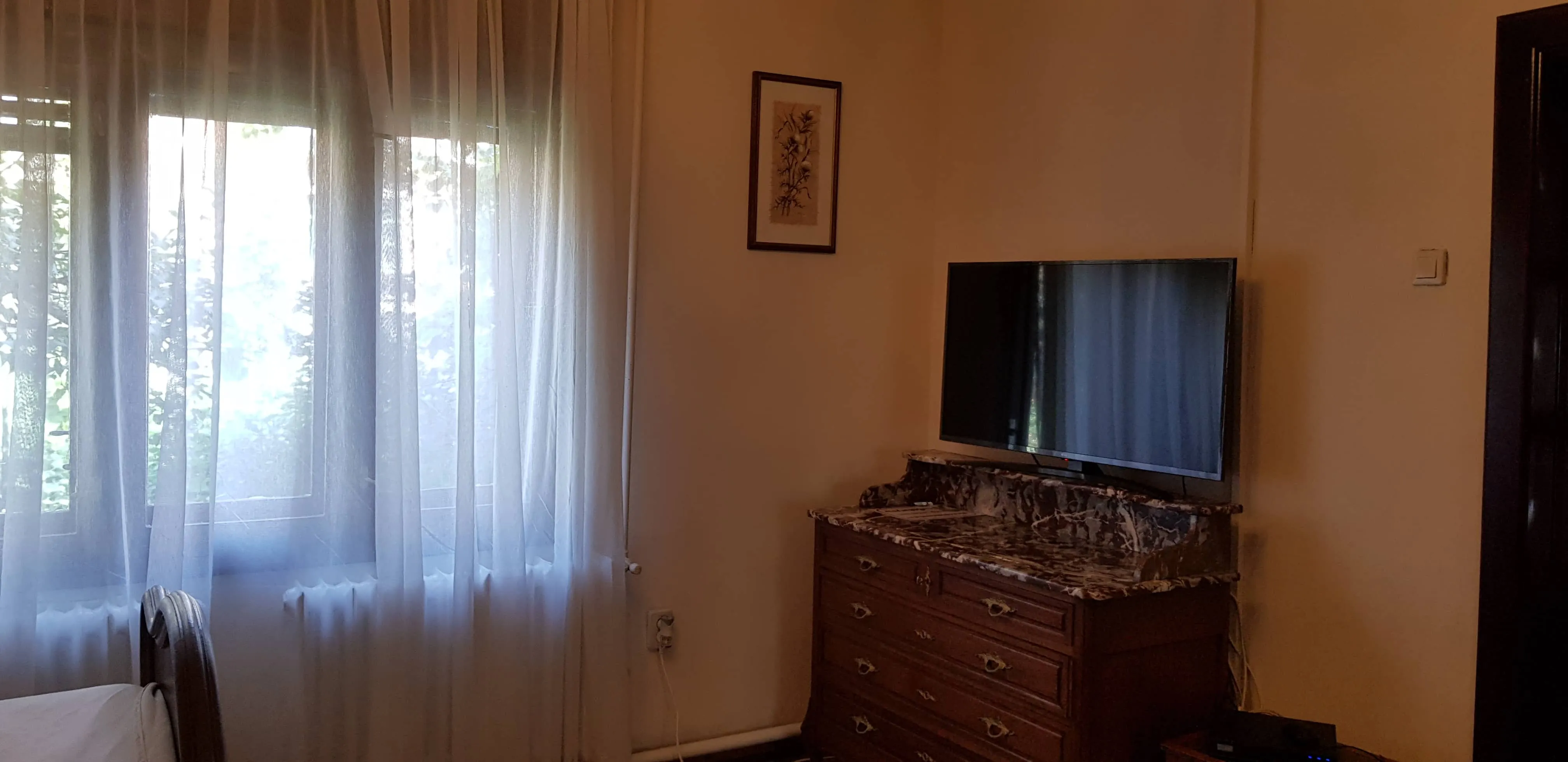 Room corner with a window covered by sheer white curtains allowing soft daylight inside, a vintage wooden dresser with a marble top holding a flat-screen TV, and a framed floral artwork on the wall above.