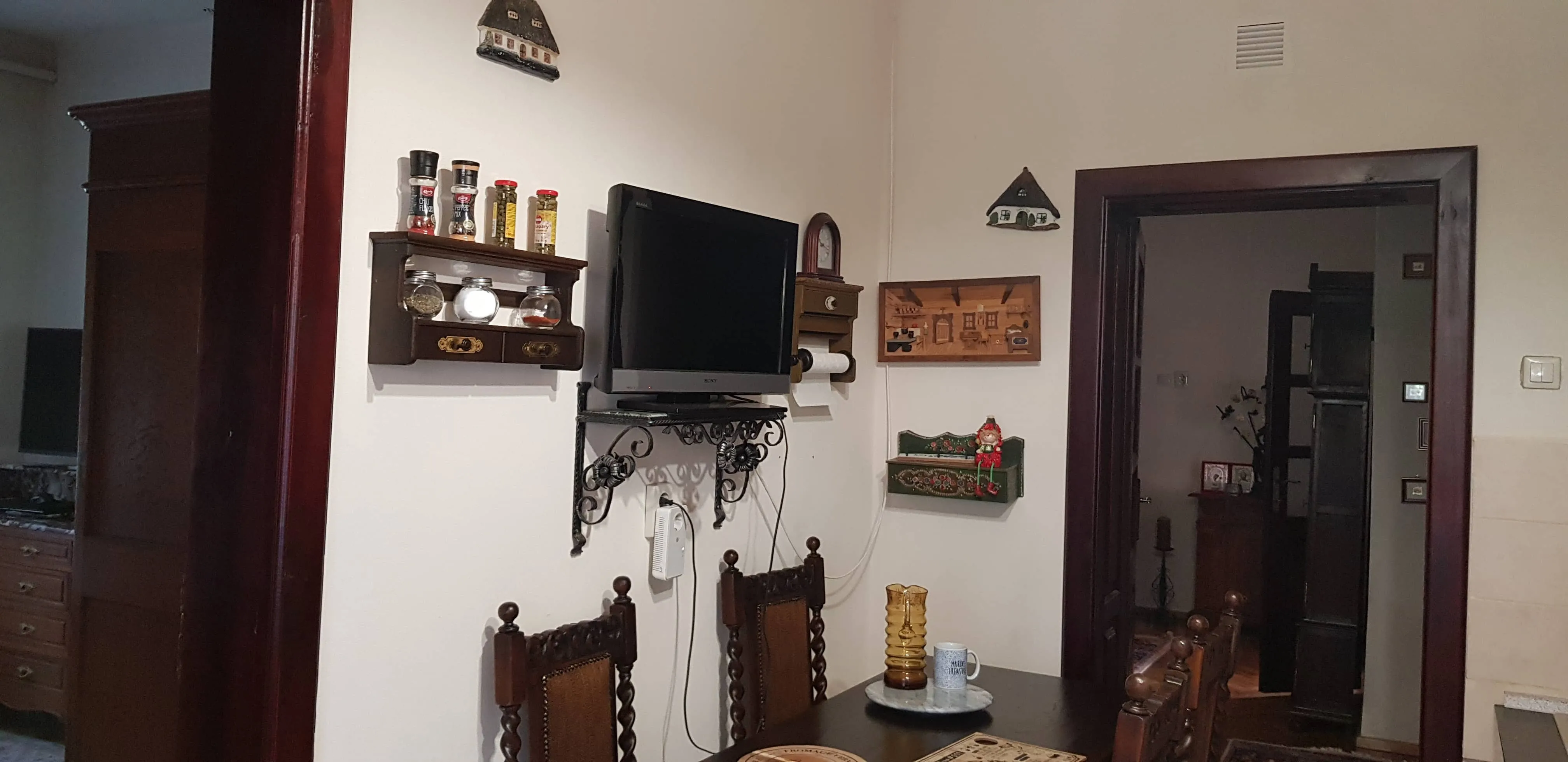 Cozy dining area with a wooden table, two ornate chairs, a mounted TV on a decorative iron shelf, spice jars on a wall shelf, and decorative plaques on a light-colored wall.