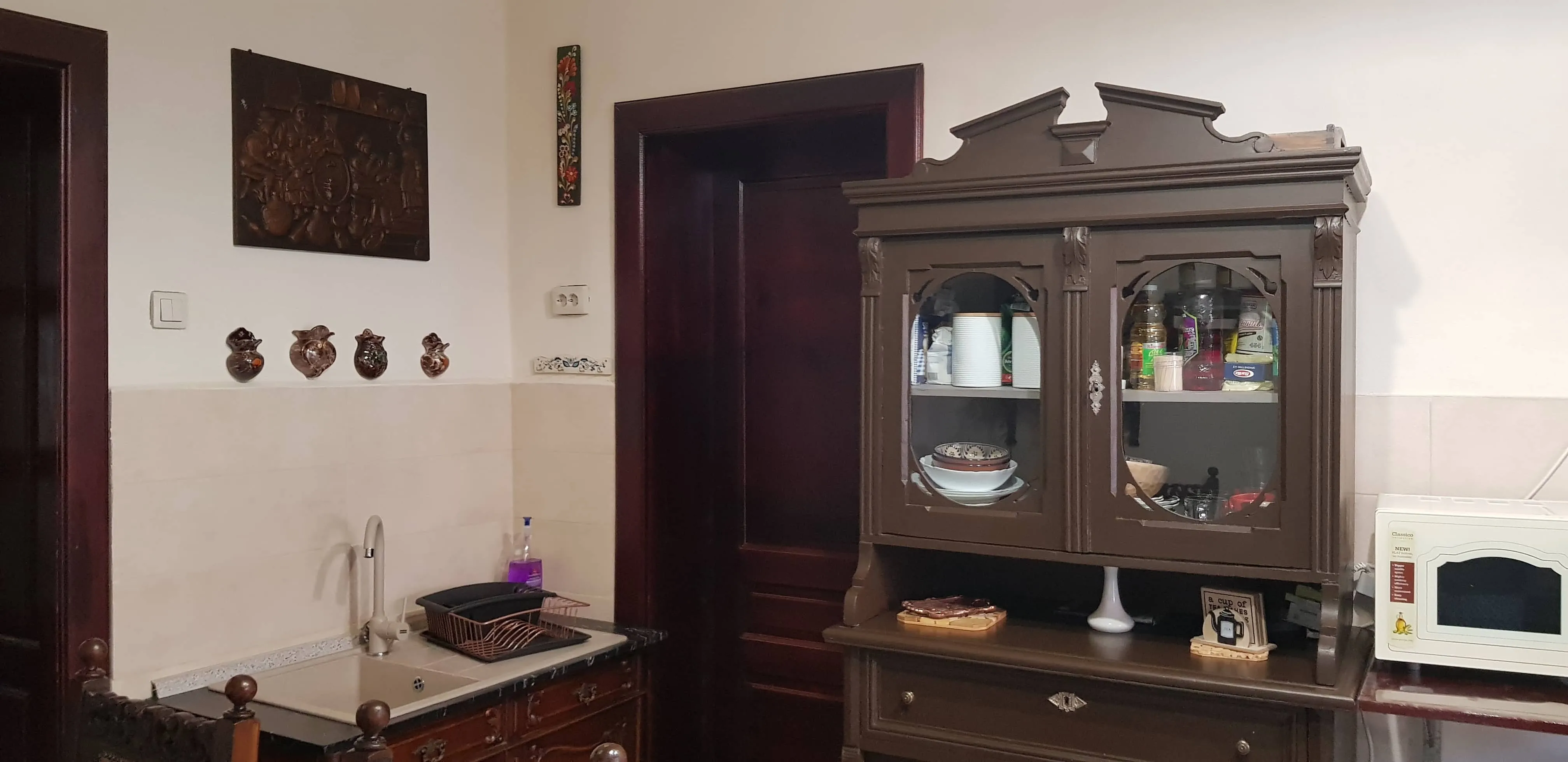 A kitchen corner featuring a dark wooden cabinet with glass doors displaying dishes and bottles, a white microwave on the right, a double sink with a faucet and a drying rack on the left, a dark doorway in the center, and decorative wall art and ceramic ornaments above the sink area.