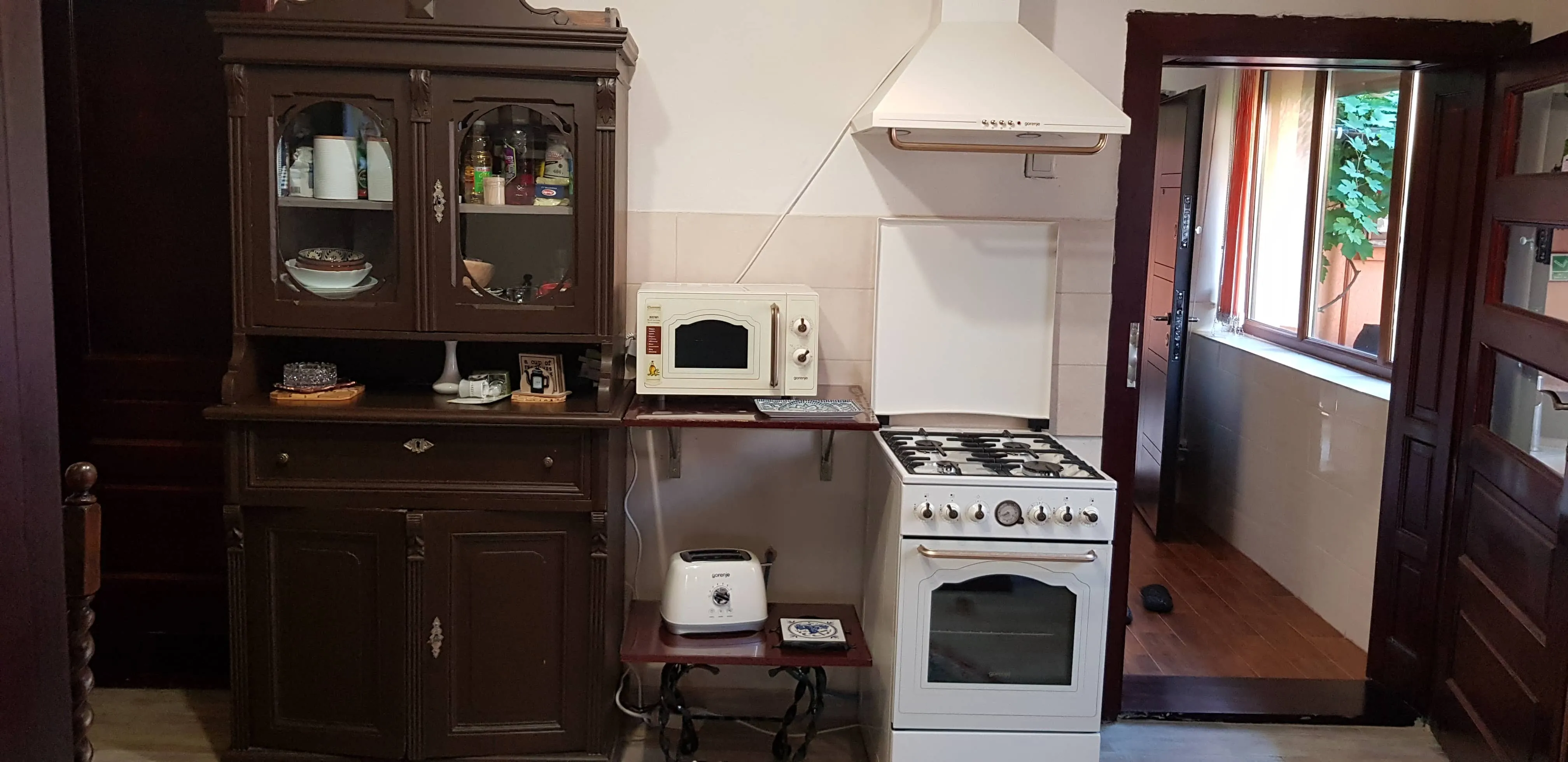 Kitchen area with a dark wooden cabinet holding bowls, bottles, and containers next to a small white microwave on a wooden shelf above a white toaster on a lower shelf. A white gas stove with six knobs and an oven is beside the shelves under a white range hood. An open doorway with a window showing green leaves is to the right.