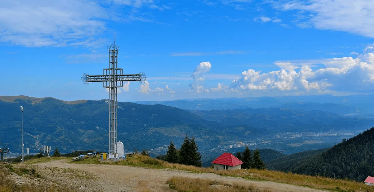 A tall metal cross structure stands on a mountain hilltop with a dirt path nearby, overlooking a vast landscape of forested hills, distant mountains, and a partly cloudy blue sky.