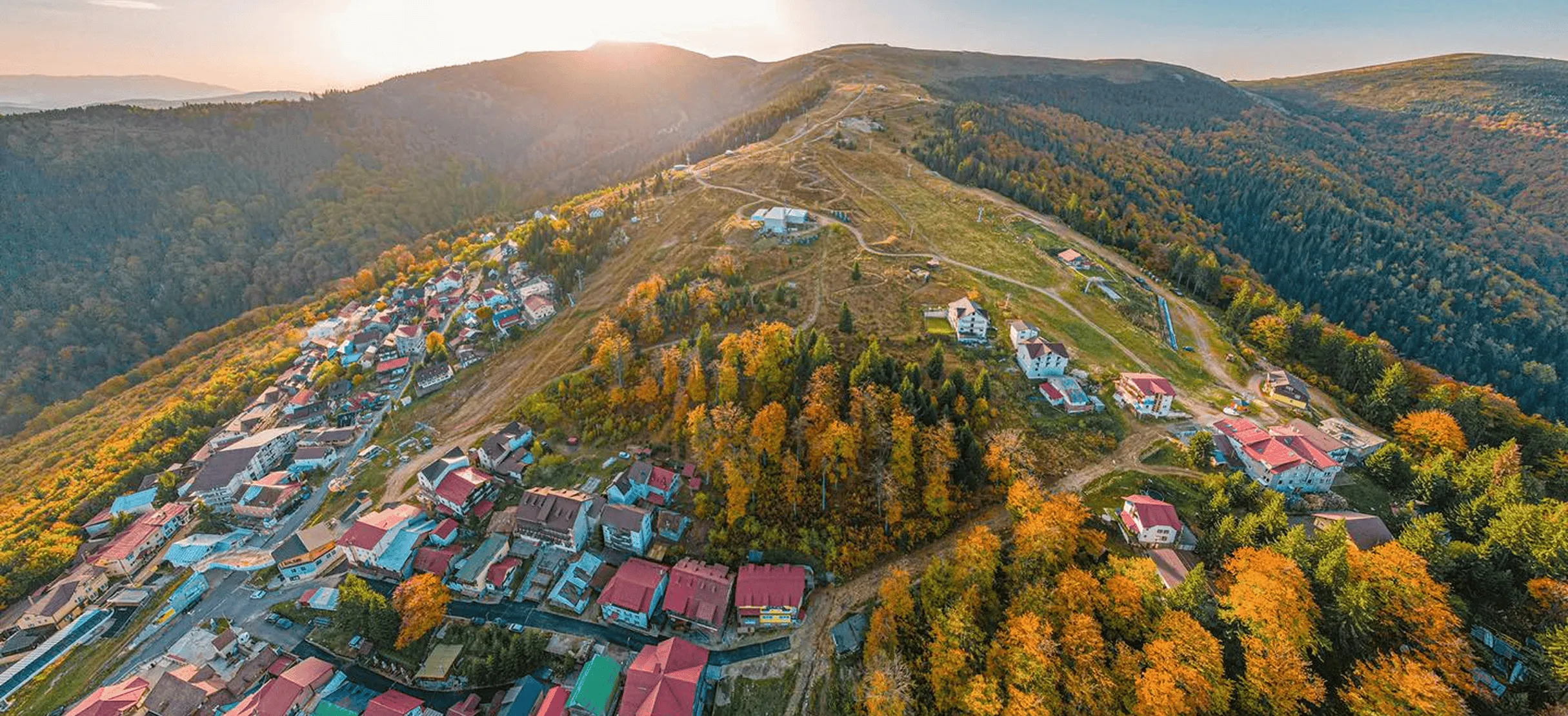 Aerial view of a village nestled on a hillside surrounded by dense forest with autumn foliage, under a setting sun behind mountains.
