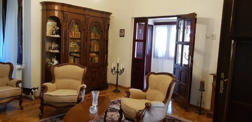 Living room with classic wooden furniture including a tall glass-fronted bookcase filled with books, two beige armchairs with carved wooden frames, an oval wooden coffee table with a glass vase, a tall black candelabra with white candles, and an open doorway with double wooden doors leading to a brightly lit adjacent room with sheer curtains. A cat is visible near a window on the left side.