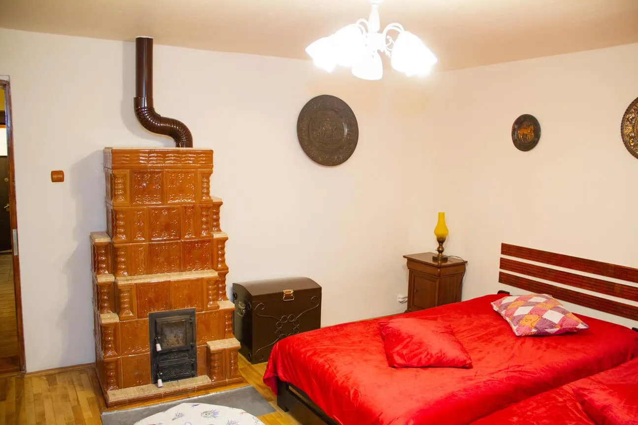 Cozy bedroom with two single beds covered in red bedspreads and pillows, a brown tiled heat stove against a white wall, wooden floor, a dark chest, a wooden nightstand with a yellow lamp, and decorative round wall hangings.