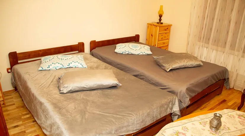 Two single wooden beds placed side by side in a room, each with a gray bedspread and two pillows, one patterned and one solid gray; a small wooden nightstand with a lamp is in the corner near sheer white curtains.