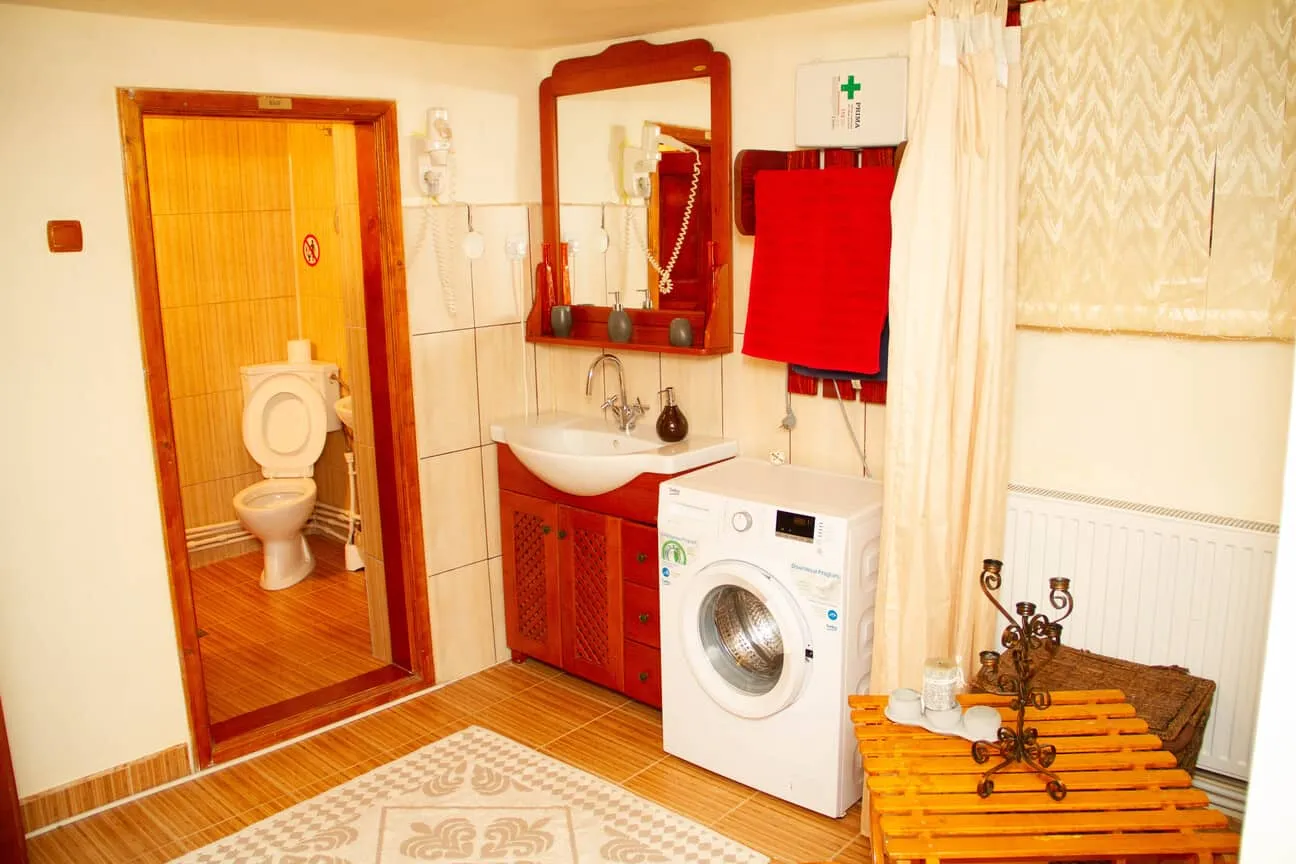 Interior of a bathroom area with a visible toilet through an open door, a sink with a wooden cabinet and mirror above it, a white front-loading washing machine, red towels hanging on the wall, a small wooden table with decorative items, and a curtained window in the background.