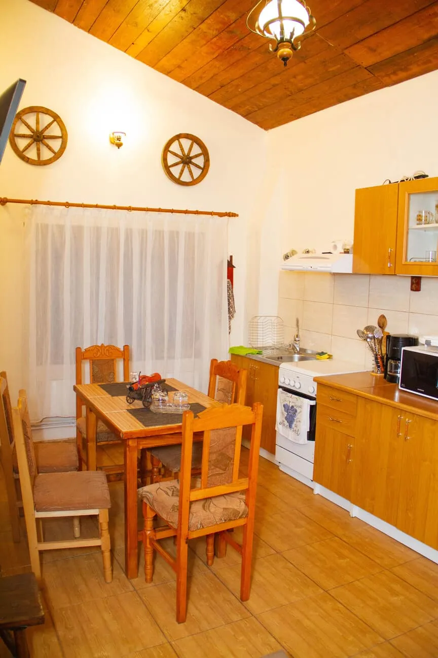Cozy kitchen with wooden ceiling and floor, featuring a wooden dining table with four cushioned chairs, a white stove with oven, a countertop with sink, a microwave, coffee maker, and utensils, and two decorative wooden wagon wheels mounted on the wall above a sheer curtain-covered window.