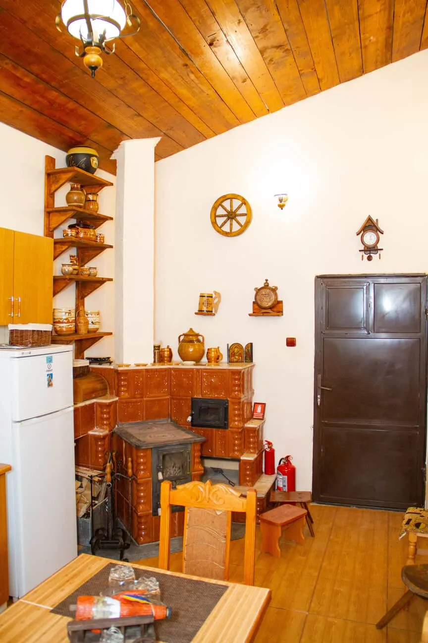 Cozy kitchen interior with wooden ceiling, tiled wood stove, white refrigerator, wooden shelves with ceramic pots, dark brown door, and wooden table with chairs.
