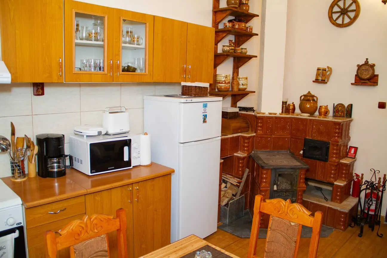 Cozy kitchen with wooden cabinets, a white refrigerator, microwave, coffee maker, toaster, paper towels, and a traditional brown tile stove with ceramic pots and wooden shelves displaying decorative items.