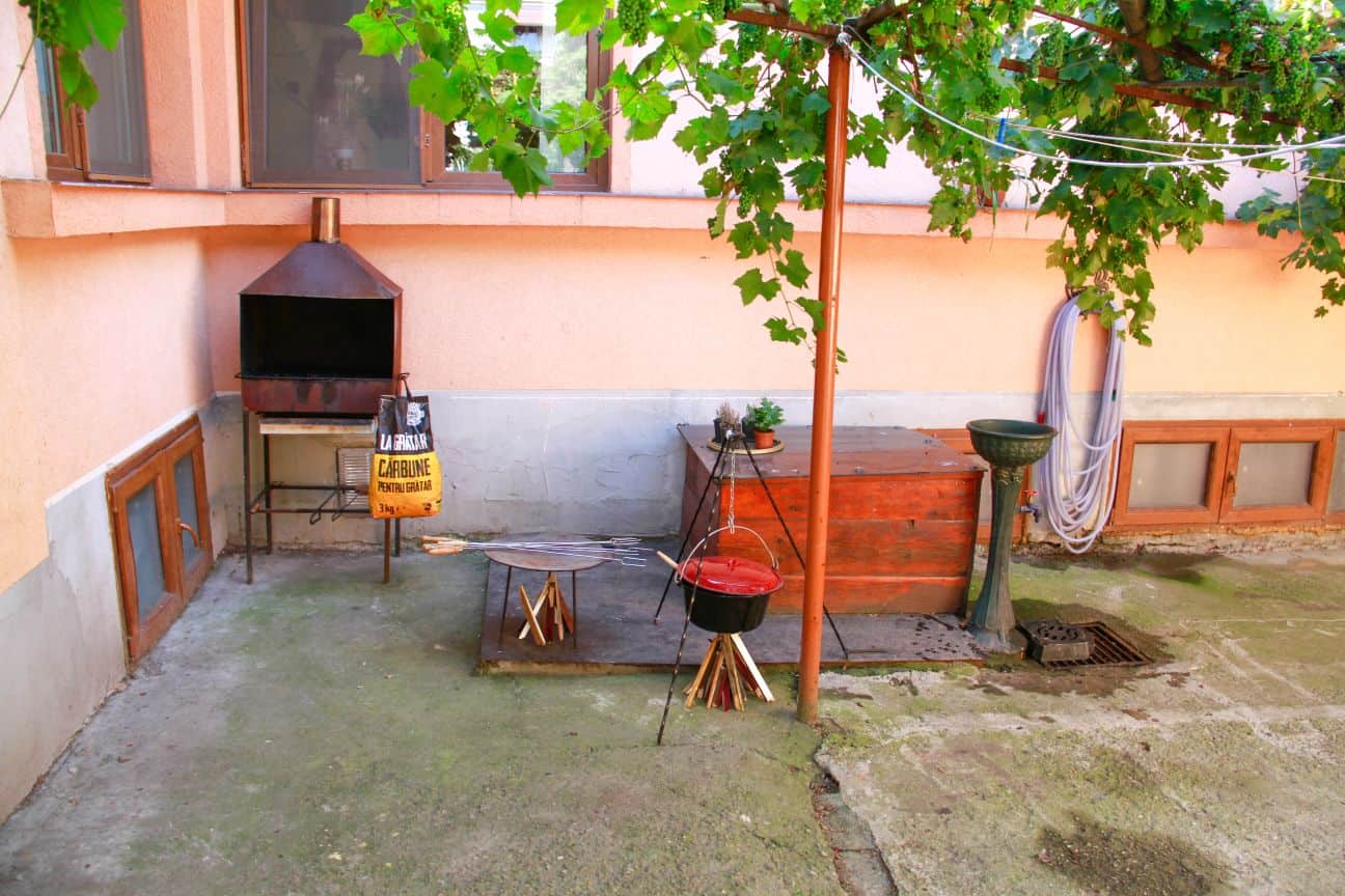 Outdoor courtyard area with a metal charcoal grill, a red cooking pot on a tripod over kindling, a round stand with skewers, a wooden storage chest, a green birdbath or fountain, a coiled hose on the wall, and vine leaves hanging from above.