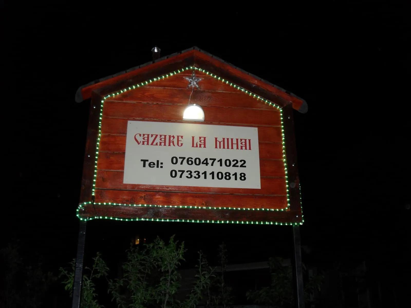 Wooden signboard with green lights outlining its shape and a star decoration at the top, displaying 'Cazare La Mihai' and two phone numbers under a small lamp at night.