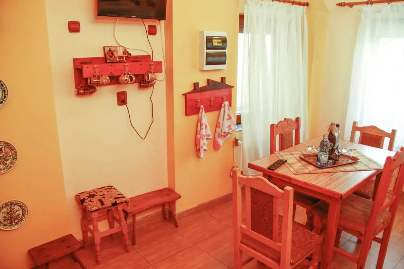 Cozy dining room with wooden furniture including a table surrounded by four carved chairs, a TV mounted on a yellow wall with ceramic mugs hanging underneath, two towels on hooks shaped like a house, and sunlight filtering through white curtains.