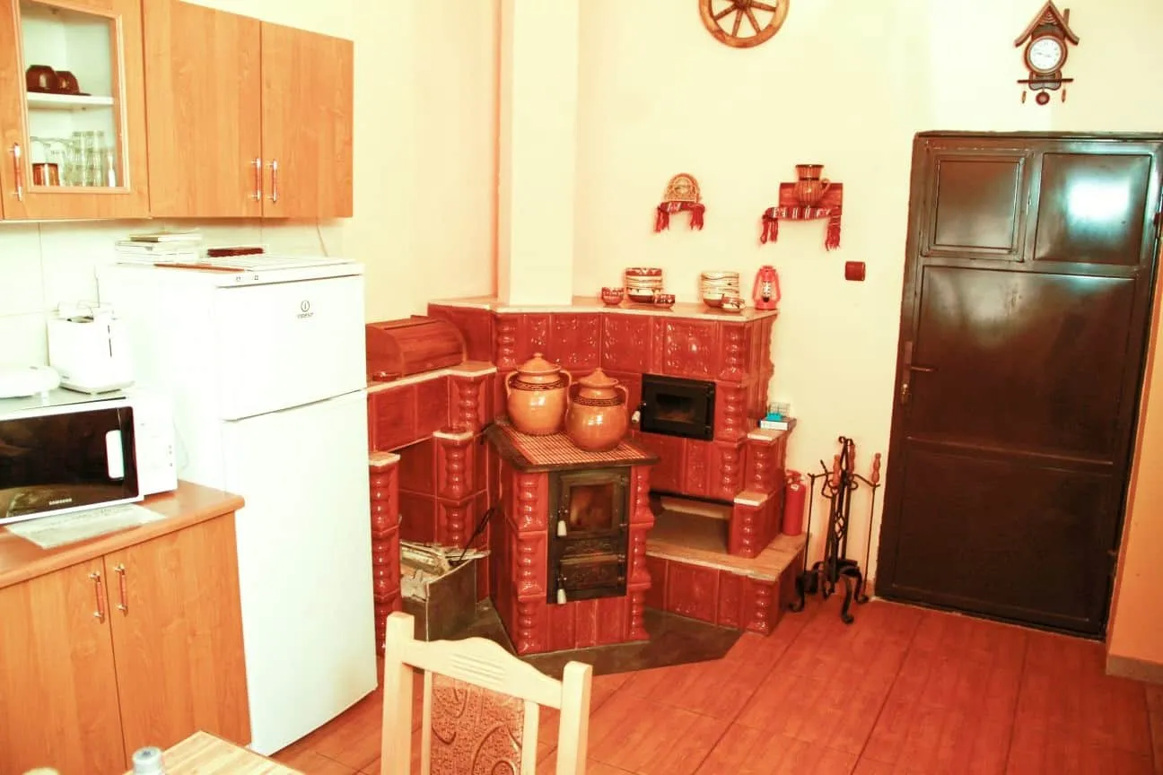 Cozy kitchen corner with a white refrigerator, microwave, wooden cabinets, and a traditional red tiled stove with brown ceramic pots on top, set against beige walls and a dark wooden door.