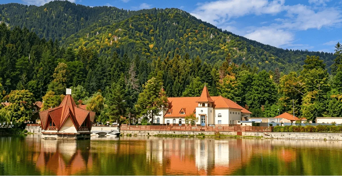 A lakeside scene featuring a white building with red roofs and a tower, next to a pavilion with multiple triangular red roofs, surrounded by greenery and forested mountains under a partly cloudy blue sky, with reflections in the calm water.