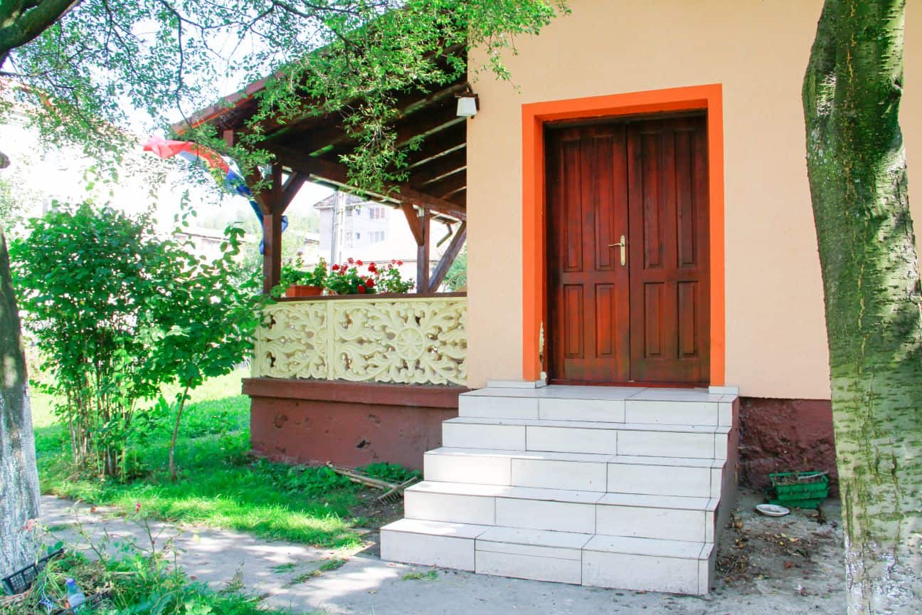 Entrance to a house with tiled steps leading up to a wooden door framed by an orange border, surrounded by green grass, trees, and a porch with decorative cream-colored railing and red flowers.