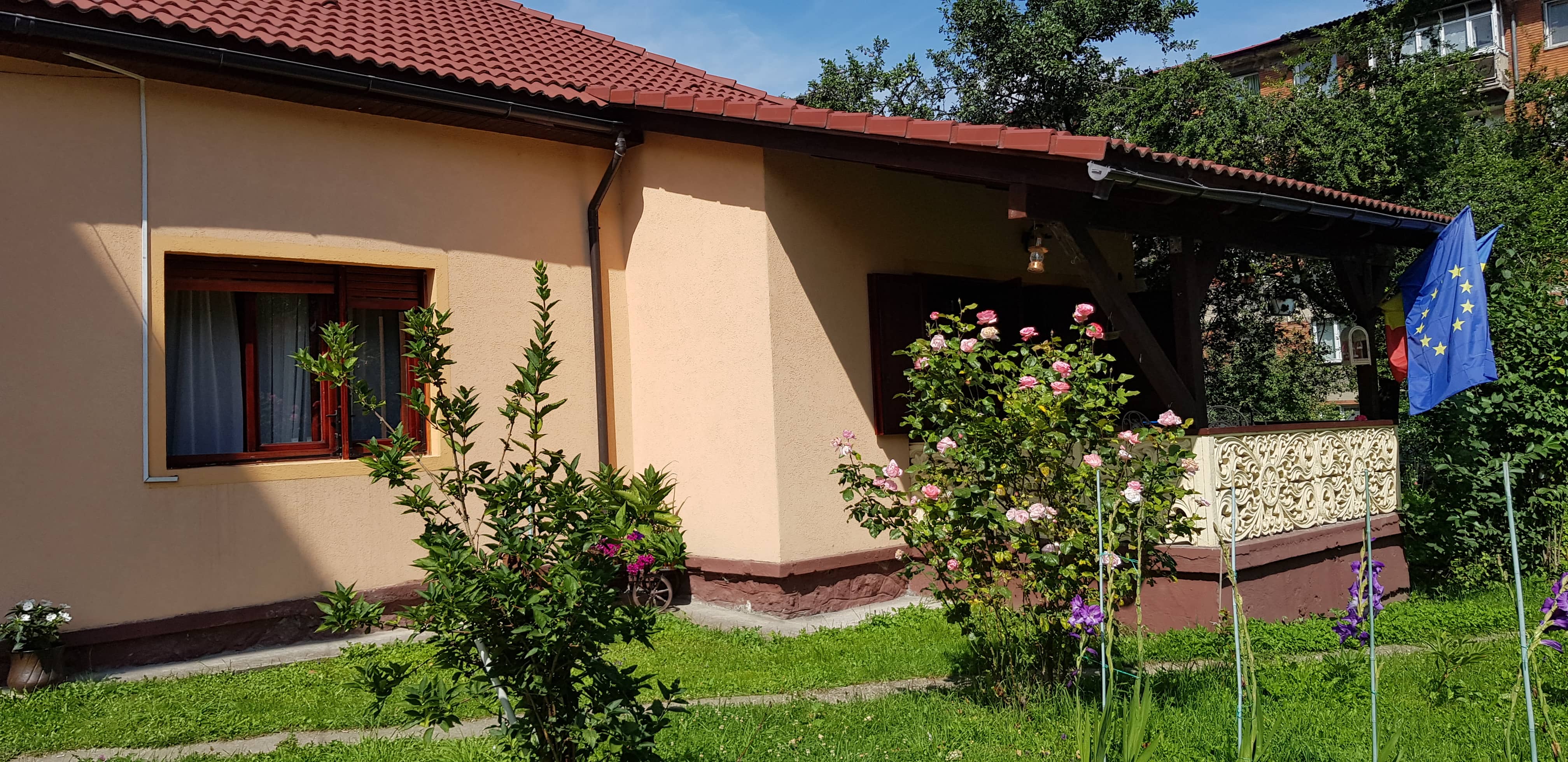 House with beige walls and red tiled roof, surrounded by a garden with green grass, blooming pink roses, purple flowers, and shrubs; balcony displays European Union and Romanian flags.