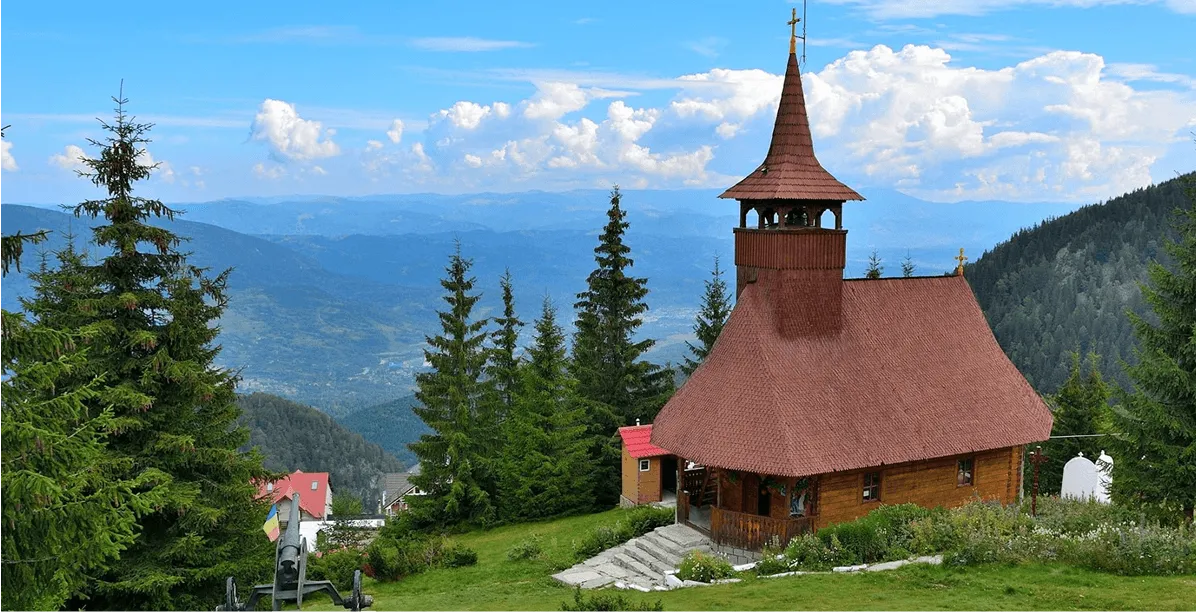 Wooden church with steep shingled roof and tall bell tower on a grassy hill surrounded by evergreen trees, with mountains and a partly cloudy blue sky in the background.