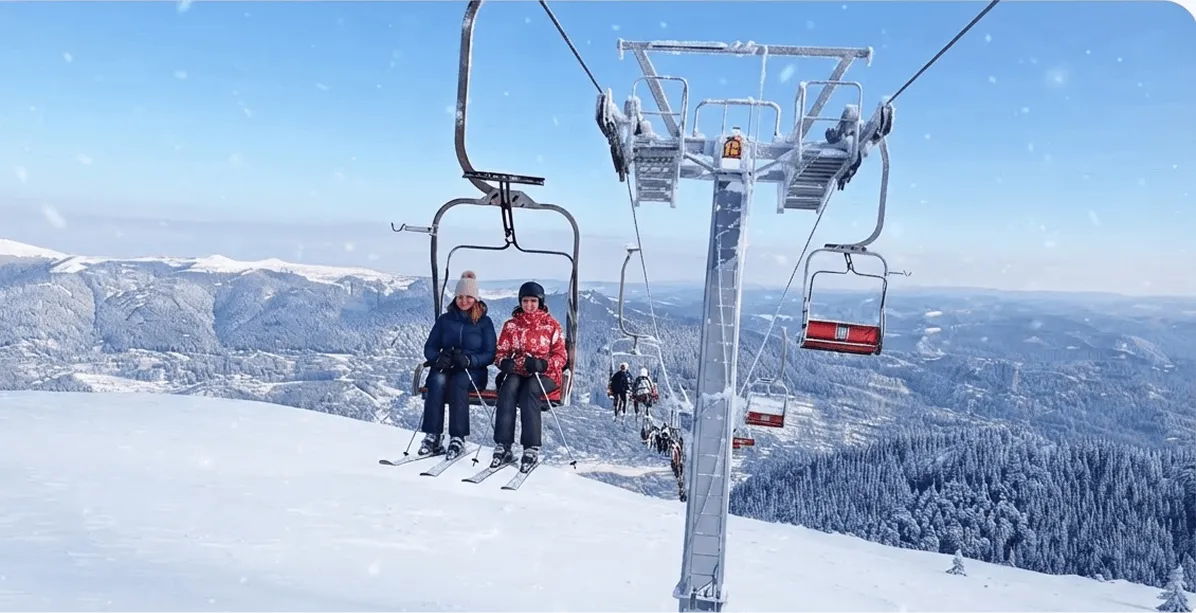 Two people wearing winter clothes and ski gear sitting on a ski lift chair, ascending over a snow-covered mountain landscape with distant snowy hills and pine trees under a clear blue sky with light snowfall.